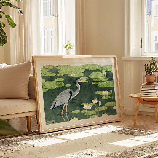 Framed art print of a blue heron standing in a lily pad pond, illustrated in a painterly watercolor style with soft greens and white lotus flowers. The artwork rests against the wall in a bright, cozy living room with beige tones, large windows, a neutral rug, and mid-century modern furniture. Sunlight streams in, casting gentle shadows and highlighting the serene nature scene.
