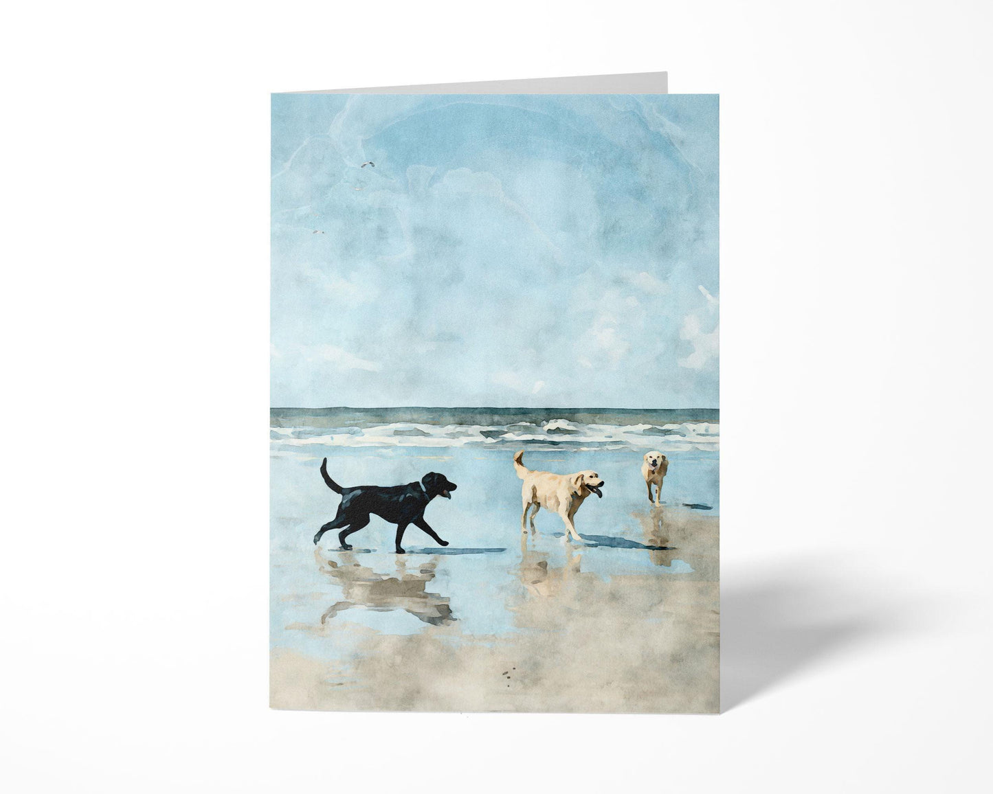 Greeting card featuring a watercolor-style illustration of three dogs—one black and two golden—playing on a sandy beach with gentle waves and a bright blue sky in the background, displayed standing upright against a white backdrop.