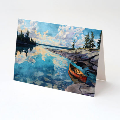 Folded greeting card featuring a watercolor lake scene with a red canoe resting on rocky shore. The calm blue water reflects soft clouds and pine trees, creating a peaceful wilderness atmosphere. Card stands open against a white background.
