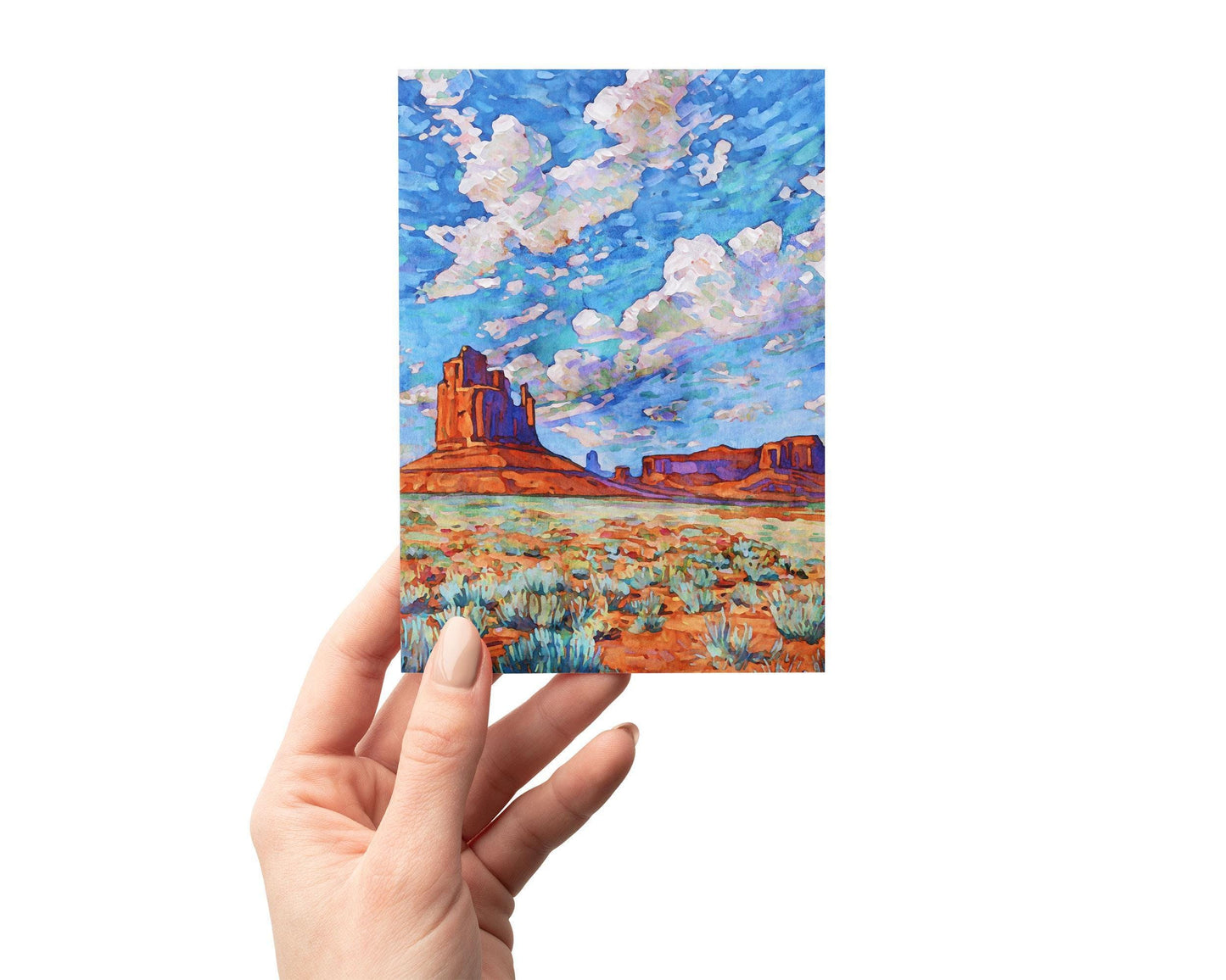 Hand holding a greeting card featuring a vivid painting of Monument Valley. The artwork shows towering red sandstone buttes rising from a desert floor with sagebrush, under a wide sky filled with swirling clouds in shades of blue, purple, and white. The impressionistic brushstrokes give the landscape a vibrant, dynamic feel, capturing the vast beauty of the American Southwest.