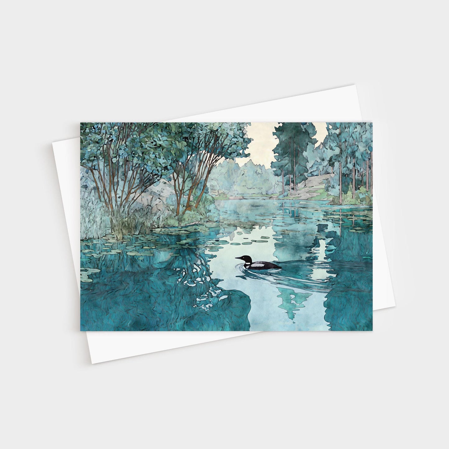 Greeting card featuring a watercolor-style illustration of a loon gliding across a calm lake. The water reflects surrounding green trees and soft blue sky, creating a serene, peaceful atmosphere. The card rests on a white envelope.