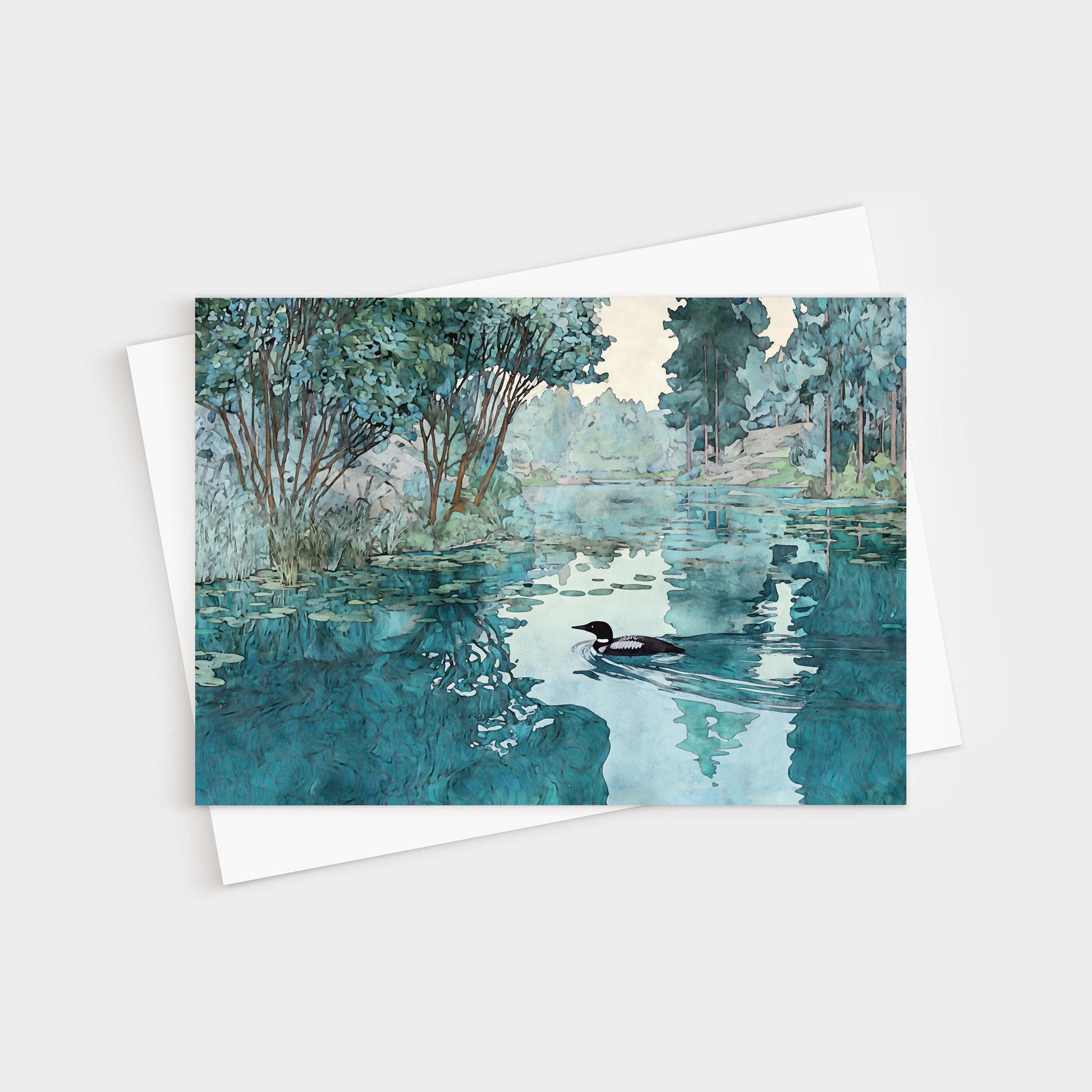 Greeting card featuring a watercolor-style illustration of a loon gliding across a calm lake. The water reflects surrounding green trees and soft blue sky, creating a serene, peaceful atmosphere. The card rests on a white envelope.
