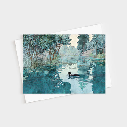 Greeting card featuring a watercolor-style illustration of a loon gliding across a calm lake. The water reflects surrounding green trees and soft blue sky, creating a serene, peaceful atmosphere. The card rests on a white envelope.