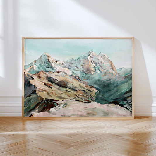Framed watercolor-style painting of snow-capped mountains in soft greens, browns, and blues, leaning against a white wall on a light wood herringbone floor with natural sunlight streaming in.