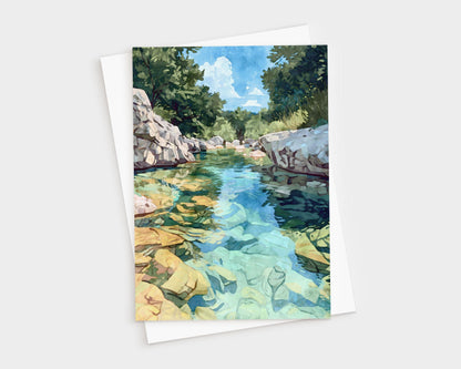 Greeting card featuring a watercolor-style painting of a crystal-clear river with golden rocks beneath the surface, bordered by white stone banks and lush green trees under a bright blue sky with clouds. Card is paired with a white envelope.