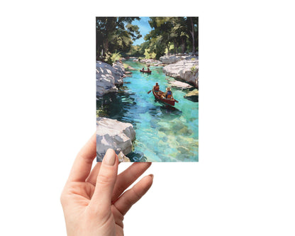 Hand holding a greeting card featuring a watercolor-style painting of two red canoes on a clear turquoise river, surrounded by white rocky banks and tall green trees under a sunny blue sky.