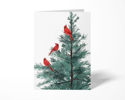 Folded greeting card featuring an illustration of three bright red cardinals perched on the branches of a green pine tree, set against a plain white background.