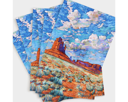 Set of greeting cards featuring a vibrant Monument Valley painting. Each card shows red rock buttes rising from a desert landscape filled with sagebrush, under a striking sky of swirling clouds in blue, purple, and white tones. The cards are fanned out in an overlapping arrangement, emphasizing the bold colors and artistic detail.