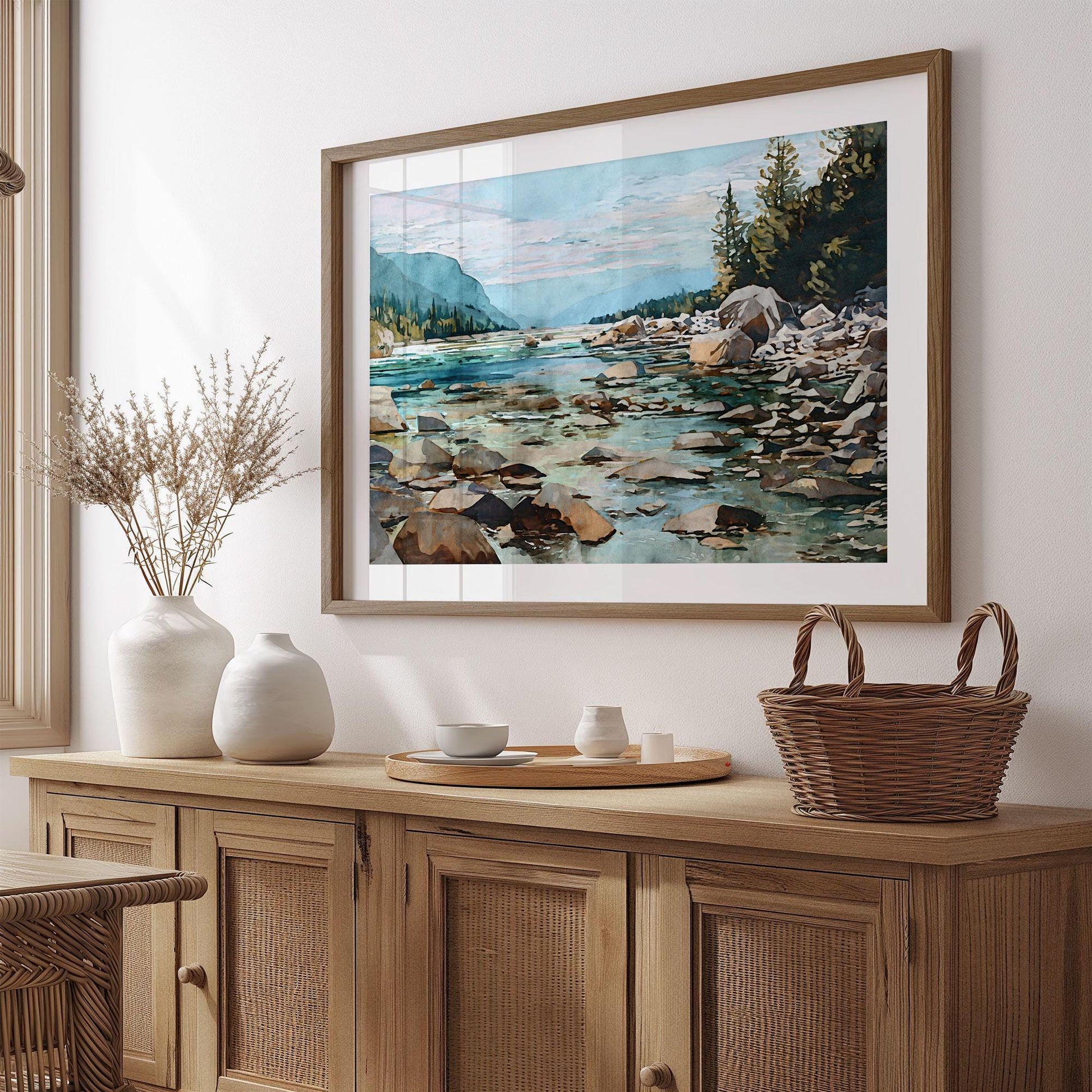 Framed watercolor landscape of a rocky river and pine forest hangs above a natural wood sideboard in a softly lit room. The painting shows turquoise water flowing past stones toward misty blue mountains under a pink-tinged sky. The decor includes white ceramic vases, a wooden tray with a candle and cup, and a woven basket, creating a warm, minimalist, nature-inspired aesthetic.
