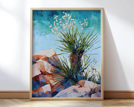 Framed painting of a desert yucca plant with spiky green leaves and white blossoms, set against angular orange and blue rock formations and a bright turquoise sky. The artwork has a modern impressionist style with bold brushstrokes and vibrant colors, leaning against a white wall on a wooden floor.