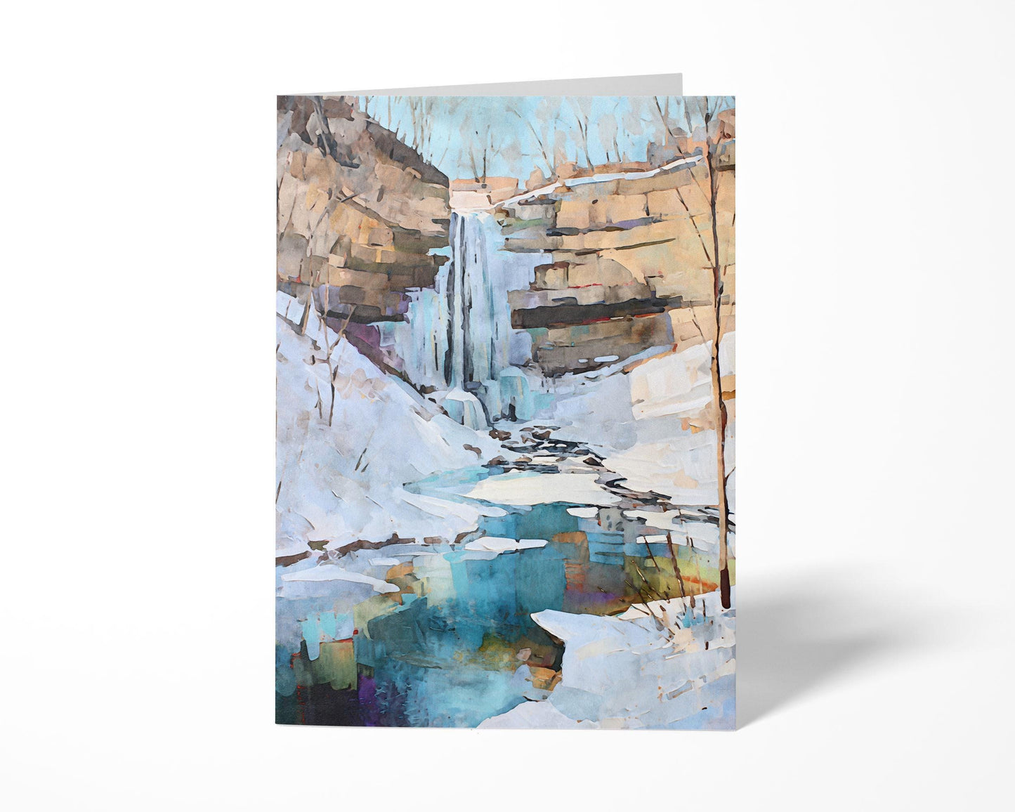 Greeting card standing upright featuring a watercolor-style painting of a snowy winter waterfall scene. The artwork shows icy cliffs, bare trees, and a turquoise pool reflecting soft tones beneath a pale blue sky.