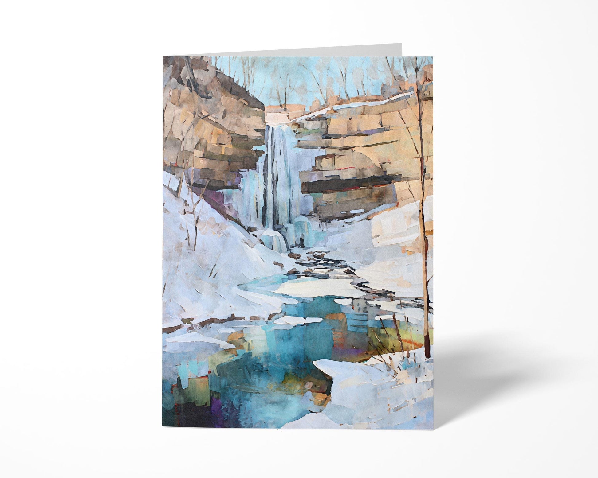Greeting card standing upright featuring a watercolor-style painting of a snowy winter waterfall scene. The artwork shows icy cliffs, bare trees, and a turquoise pool reflecting soft tones beneath a pale blue sky.