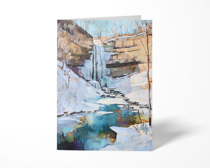 Greeting card standing upright featuring a watercolor-style painting of a snowy winter waterfall scene. The artwork shows icy cliffs, bare trees, and a turquoise pool reflecting soft tones beneath a pale blue sky.