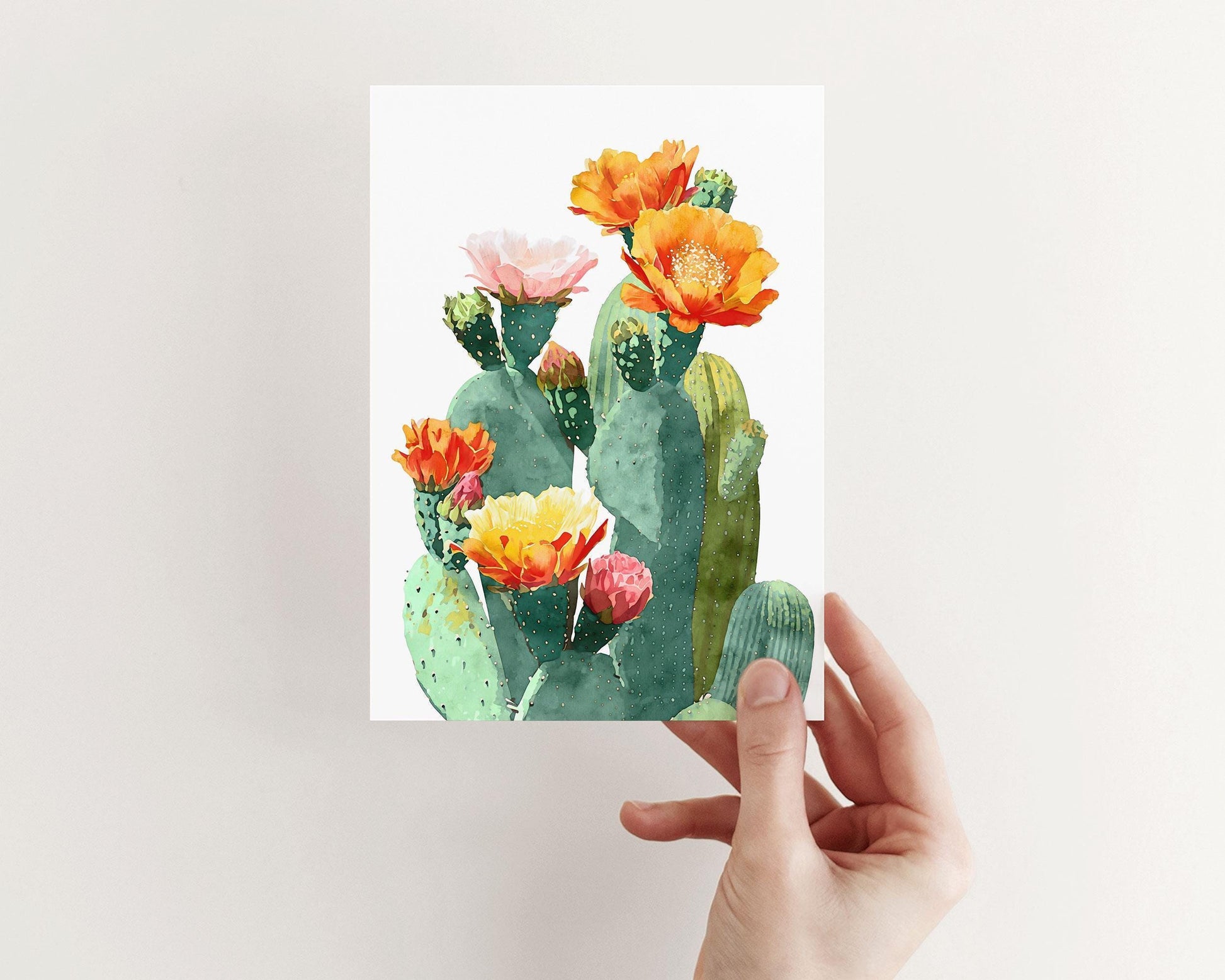 Hand holding a greeting card with a watercolor illustration of green prickly pear cacti topped with colorful blossoms in orange, yellow, pink, and red, set against a clean white background.