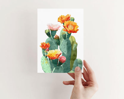 Hand holding a greeting card with a watercolor illustration of green prickly pear cacti topped with colorful blossoms in orange, yellow, pink, and red, set against a clean white background.