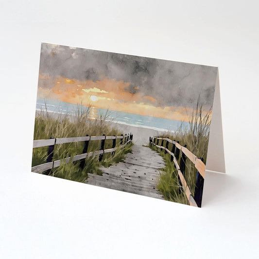 Greeting card featuring a watercolor-style illustration of a wooden boardwalk lined with weathered rails, leading through beach grass to a serene ocean sunset under moody gray skies, shown upright on a white background.