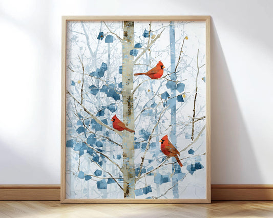 Framed art print of three bright red cardinals perched on slender birch branches in a winter forest. Soft blue leaves and abstract geometric foliage create a cool, snowy backdrop. The natural wood frame rests against a white wall on a light wood floor, highlighting the contrast between the vivid birds and the muted winter tones.