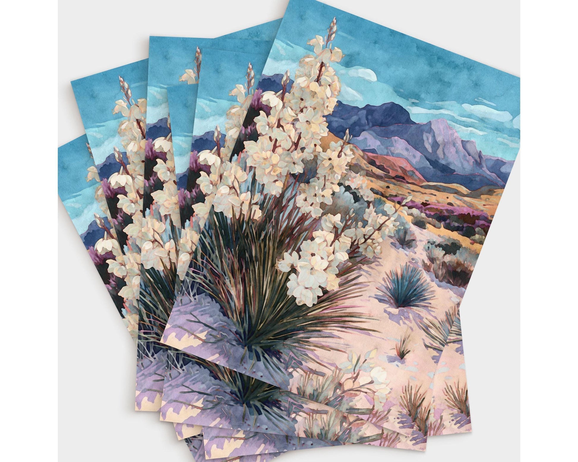 Set of greeting cards arranged in a fanned stack, each featuring a watercolor desert landscape with blooming yucca plants in the foreground, sandy terrain, and purple mountains under a bright blue sky with soft clouds.