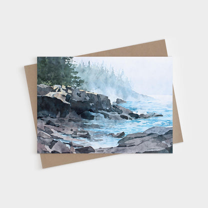 Horizontal greeting card featuring a watercolor-style illustration of a misty Lake Superior shoreline. Dark, layered rocky cliffs extend into calm blue water, with gentle waves lapping against the stones. A soft fog drifts across the scene, partially obscuring a distant forest of evergreen trees. Muted blues, grays, and greens create a quiet, atmospheric coastal mood.