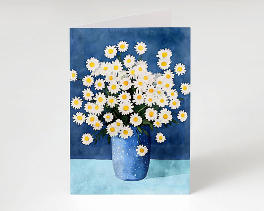 Folded greeting card standing upright, featuring a watercolor illustration of white daisies with yellow centers in a blue polka dot vase, set against a dark blue background and light blue tabletop.