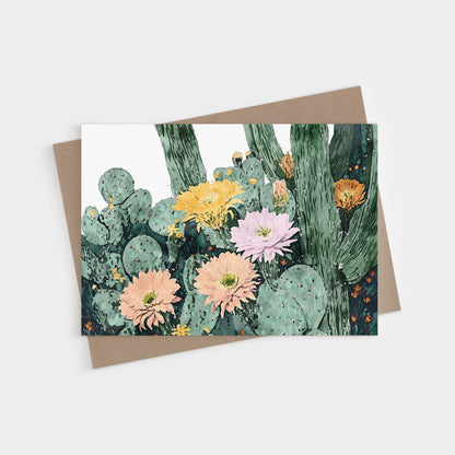 Greeting card featuring a watercolor illustration of blooming cacti with large flowers in soft peach, pink, yellow, and orange tones against green cactus pads and tall stems, paired with a natural brown envelope.