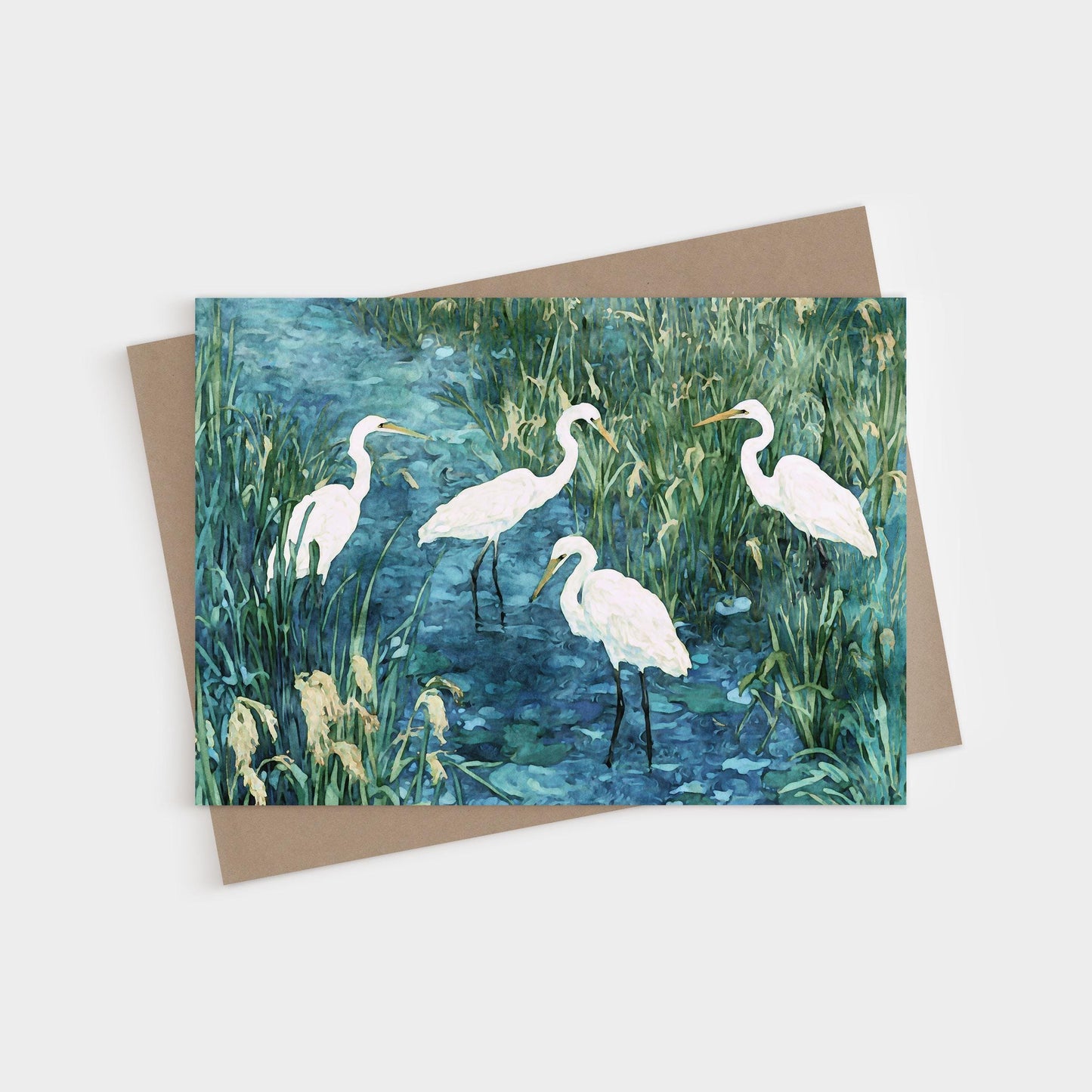 Greeting card featuring a watercolor-style painting of five white egrets standing in a marshy blue-green wetland, surrounded by tall grasses and reeds. Card is paired with a kraft envelope.