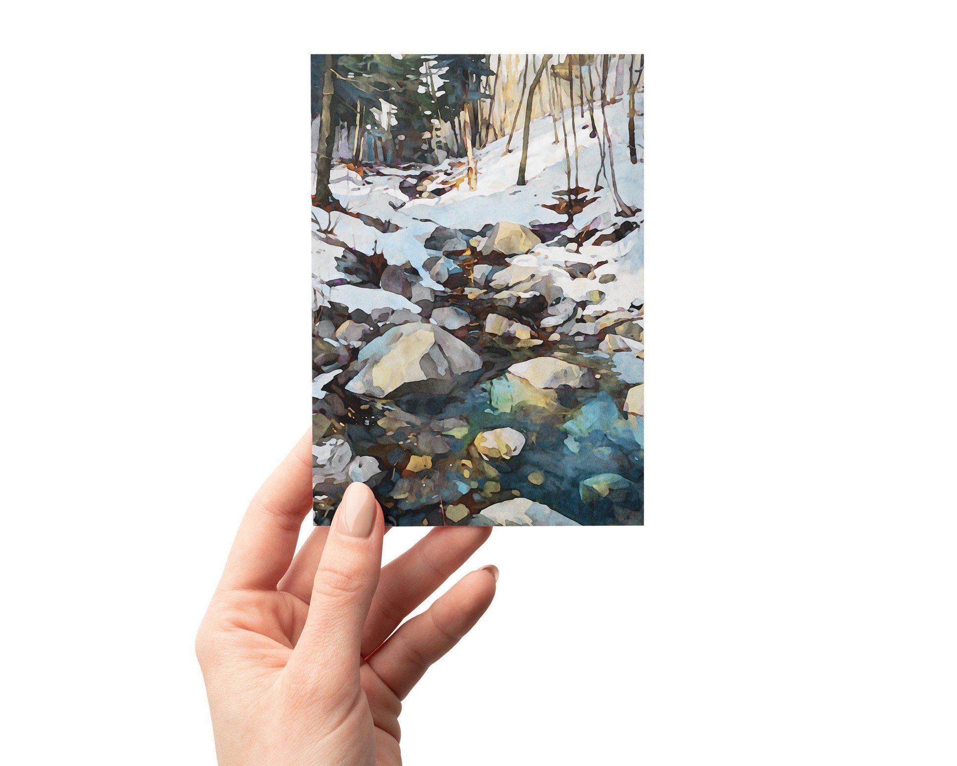 Hand holding a greeting card featuring a watercolor-style winter landscape of a rocky creek flowing through snow-covered woods, with sunlight reflecting on the icy blue and green water.