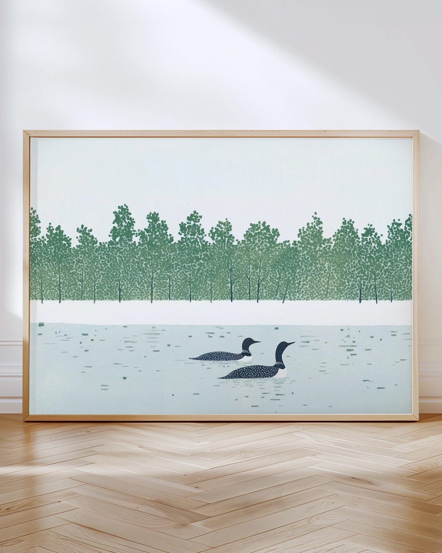 Framed art print of two loons swimming in a serene lake with a dense green tree line in the background. The artwork leans against a white wall on a light wood herringbone floor, bathed in natural sunlight for a peaceful, modern display.