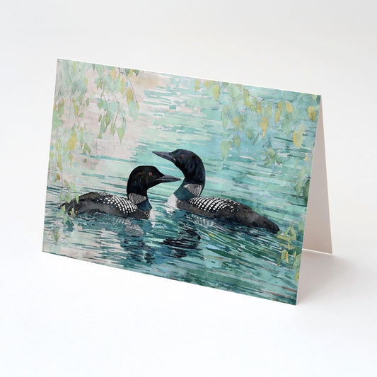 Folded watercolor greeting card featuring two loons gliding side by side on a serene turquoise lake with leafy branches overhead, displayed upright against a white background.