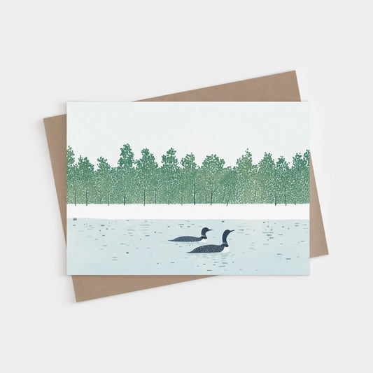 Greeting card featuring a minimalist illustration of two loons swimming on a calm lake, with a dense green treeline in the background. The artwork uses soft, muted tones of blue, green, and white. A kraft brown envelope is partially visible beneath the card.