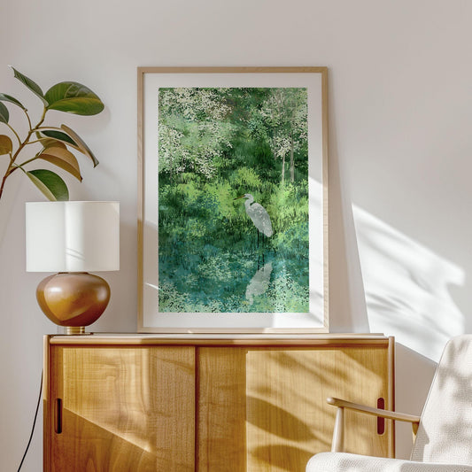 Framed watercolor-style art print featuring a white egret by a reflective pond, surrounded by lush green grasses and blossoming trees. Displayed in a sunlit modern room on a wooden cabinet beside a ceramic lamp and leafy plant, creating a calm, nature-inspired interior.