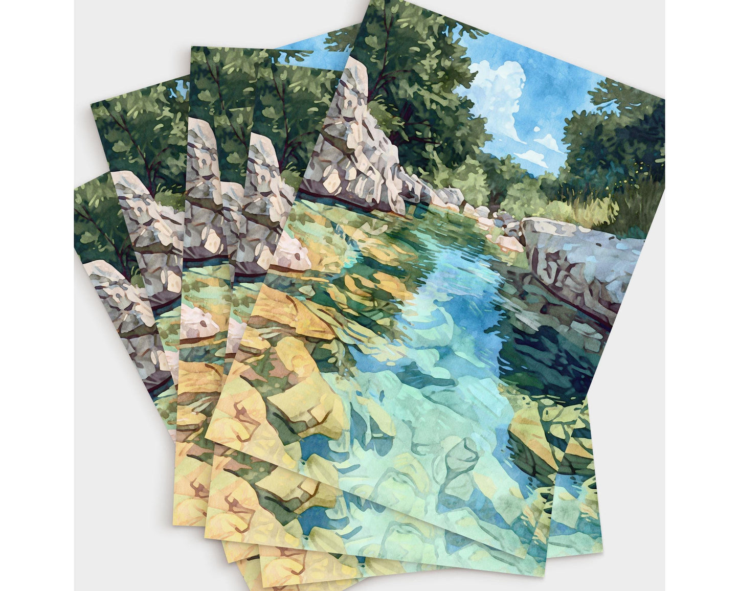 Set of greeting cards featuring a watercolor-style painting of a turquoise river with golden rocks beneath the water, framed by rocky white banks and lush green trees under a bright blue sky. Cards are fanned out in a layered display.