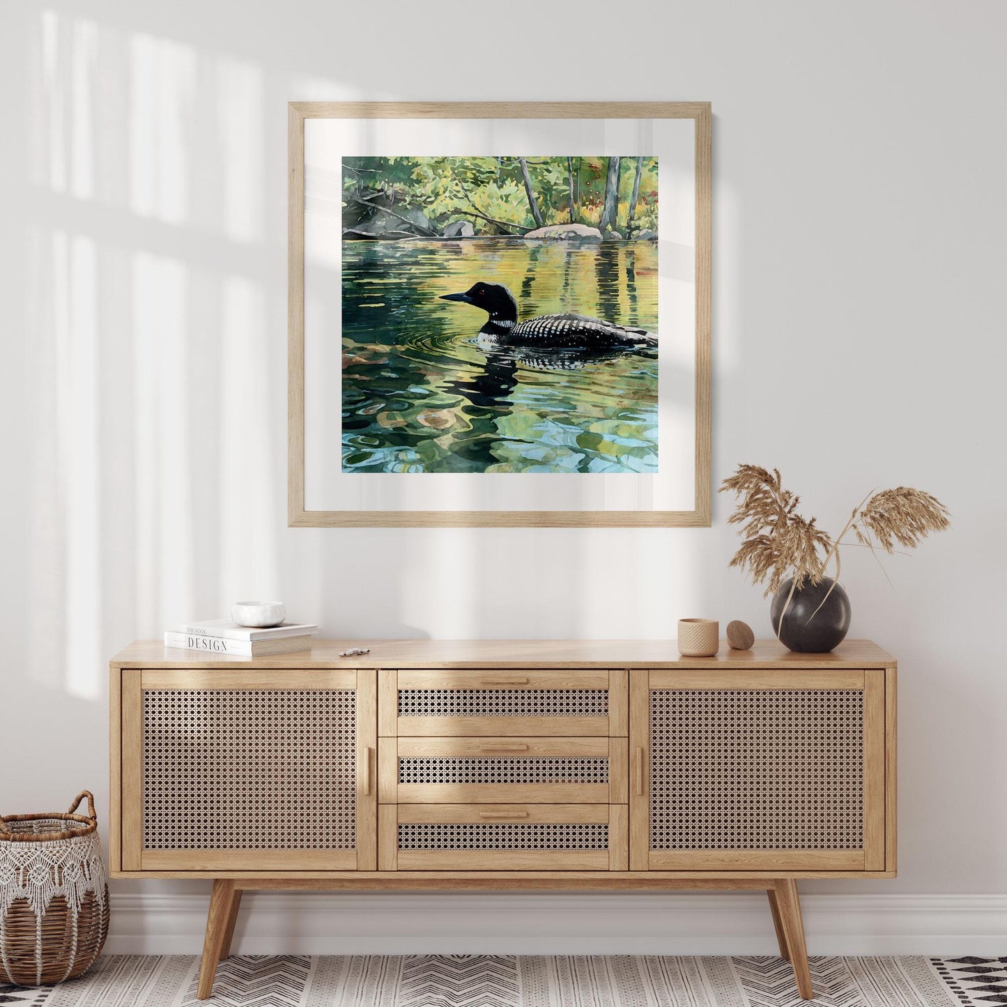 Framed art print of a loon gliding across a sunlit lake, its black-and-white plumage mirrored in shimmering green and gold reflections. The artwork hangs above a mid-century cane sideboard styled with a black vase of dried grasses, neutral ceramics, and books. Soft morning light filters through the room, creating a warm, natural, Scandinavian-inspired setting.