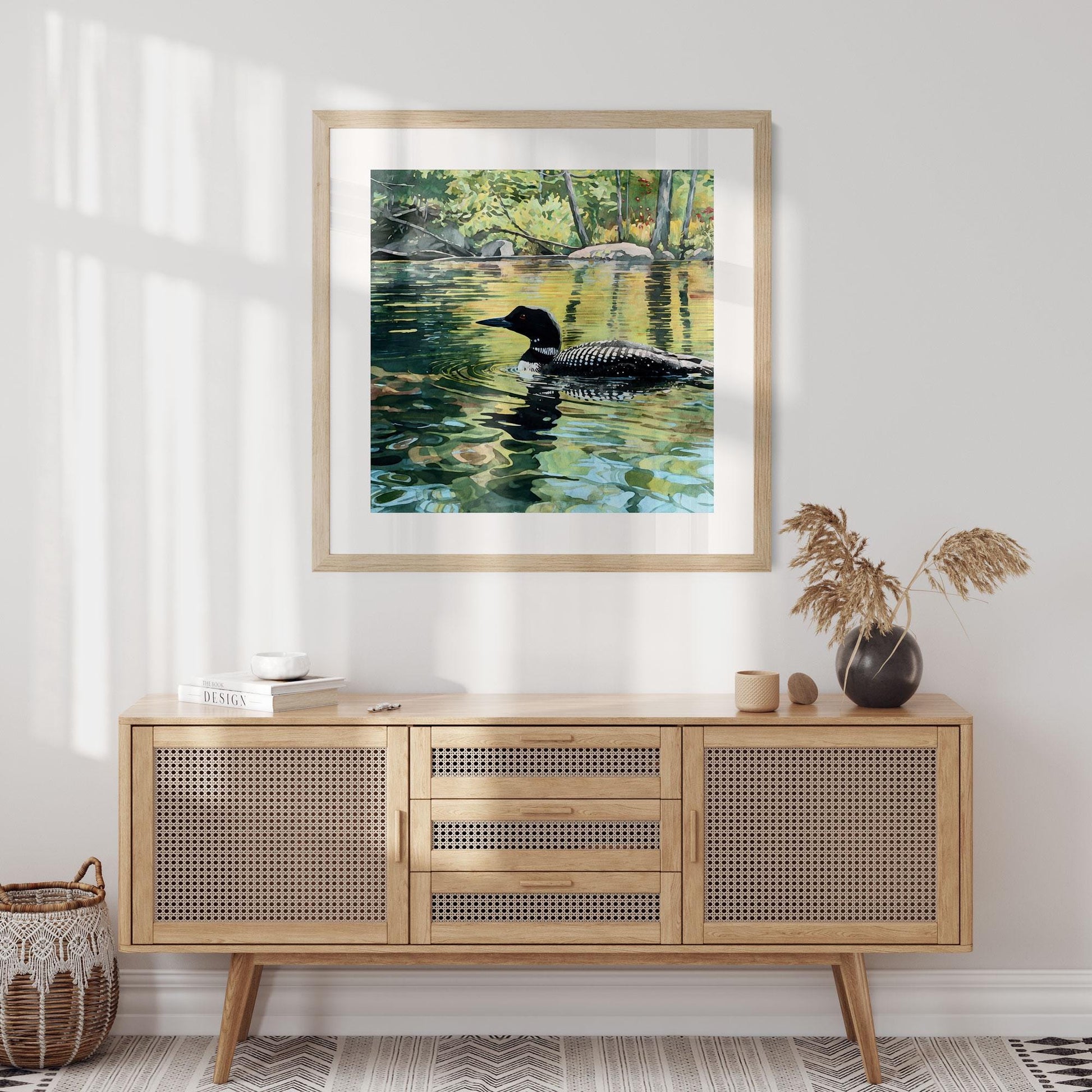 Framed art print of a loon gliding across a sunlit lake, its black-and-white plumage mirrored in shimmering green and gold reflections. The artwork hangs above a mid-century cane sideboard styled with a black vase of dried grasses, neutral ceramics, and books. Soft morning light filters through the room, creating a warm, natural, Scandinavian-inspired setting.