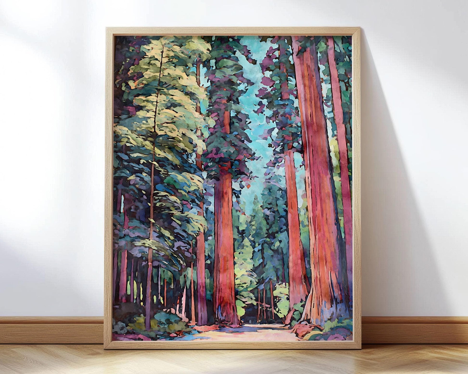 Framed art print of a colorful impressionist-style forest scene, featuring tall redwood trees with reddish bark and deep green foliage, against a bright sky in shades of blue and turquoise. The artwork rests on a light wooden floor, leaning against a white wall.
