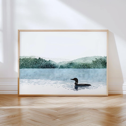 Minimalist art print featuring a loon swimming in a calm blue lake with rippling water, set against layered green forested hills and a pale sky, framed in light wood and displayed on a floor with herringbone wood flooring.