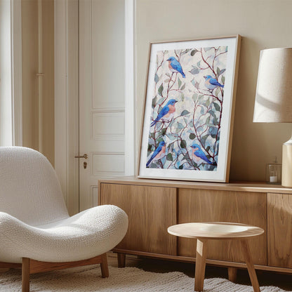 Framed watercolor-style print of bluebirds perched among leafy branches, displayed on a wooden credenza in a softly lit modern living room. The artwork features blue and peach birds with green and teal foliage on a pale background. The space includes a curved white boucle chair, light wood furniture, a neutral rug, and a beige wall, creating a warm, minimalist, Scandinavian-inspired setting.