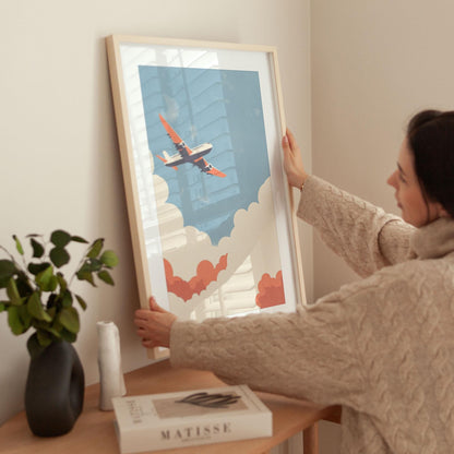 Woman in a cozy beige sweater places a framed art print of an airplane with orange wings flying through stylized clouds against a blue sky on a wooden table; nearby are a black vase with greenery, a sculptural white object, and a Matisse art book.