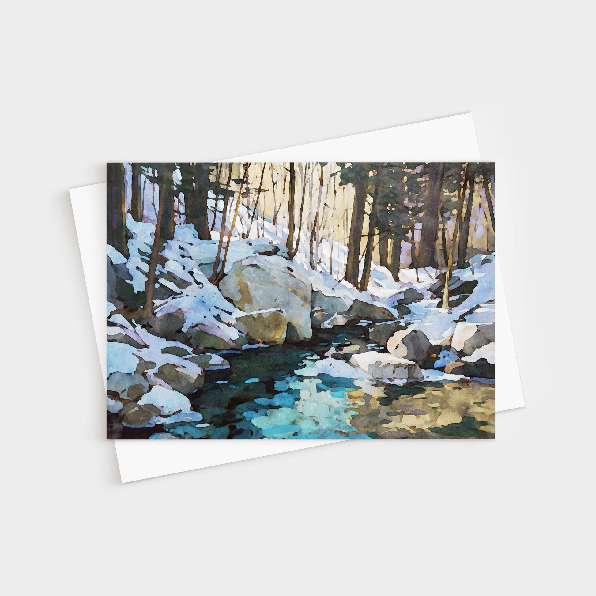 Greeting card featuring a watercolor-style painting of a snowy forest scene with large rocks and a creek reflecting icy blue and golden tones. The card is displayed with a white envelope underneath.