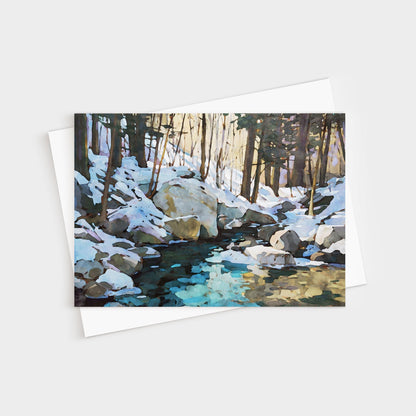 Greeting card featuring a watercolor-style painting of a snowy forest scene with large rocks and a creek reflecting icy blue and golden tones. The card is displayed with a white envelope underneath.