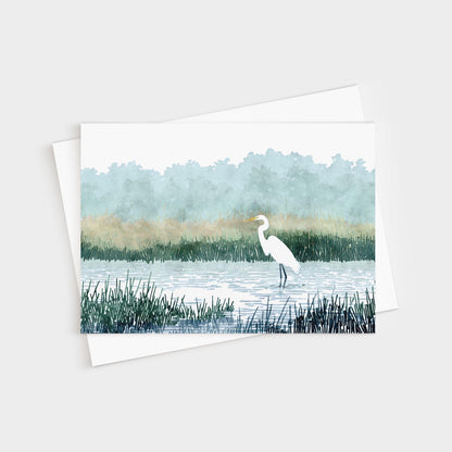Watercolor-style illustration of a white heron standing in a marsh, surrounded by tall grasses and calm water, with soft green trees and golden reeds in the misty background. The card is displayed with a white envelope.