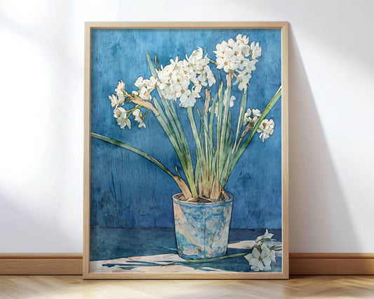 Framed art print of white narcissus flowers in a mottled blue ceramic pot, painted in a watercolor style. The blossoms stand tall with long green leaves against a textured deep blue background. One stem with flowers lies at the base on a white cloth, adding a natural touch to the minimalist composition. The frame rests on a light wooden floor, leaning casually against a wall.