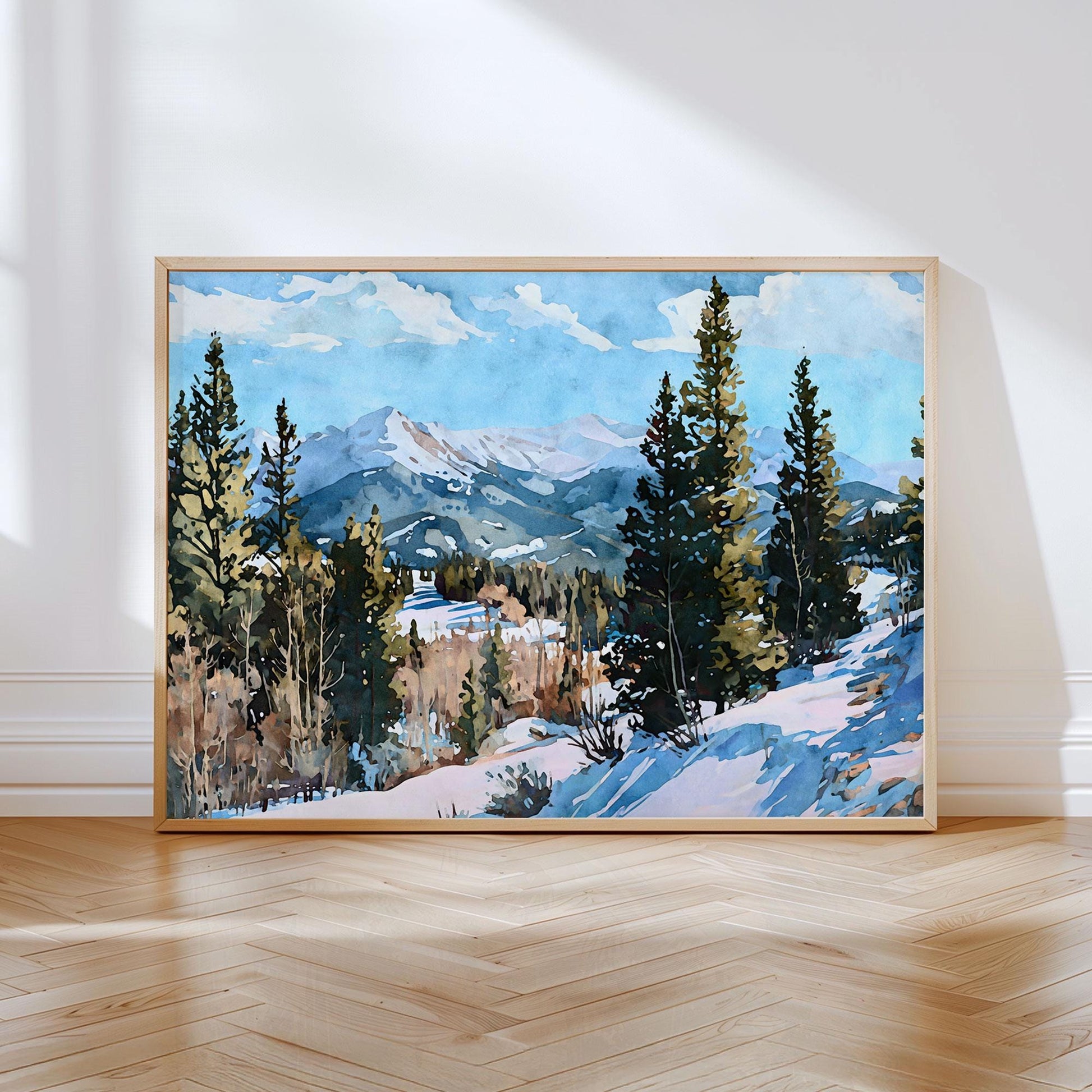 Watercolor landscape painting of a snow-covered mountain range framed by evergreen trees under a bright blue sky with scattered clouds, displayed in a light wood frame leaning against a white wall on a herringbone wood floor.