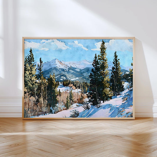 Watercolor landscape painting of a snow-covered mountain range framed by evergreen trees under a bright blue sky with scattered clouds, displayed in a light wood frame leaning against a white wall on a herringbone wood floor.