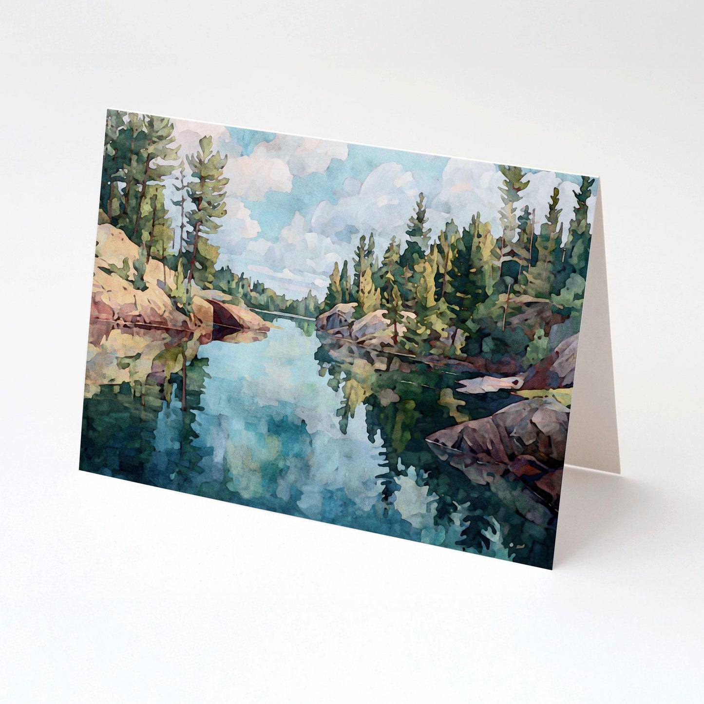 Folded greeting card featuring a watercolor-style painting of a peaceful northern lake, with rocky shores and tall evergreen trees reflected in the calm blue water under a partly cloudy sky. Displayed upright on a white background.