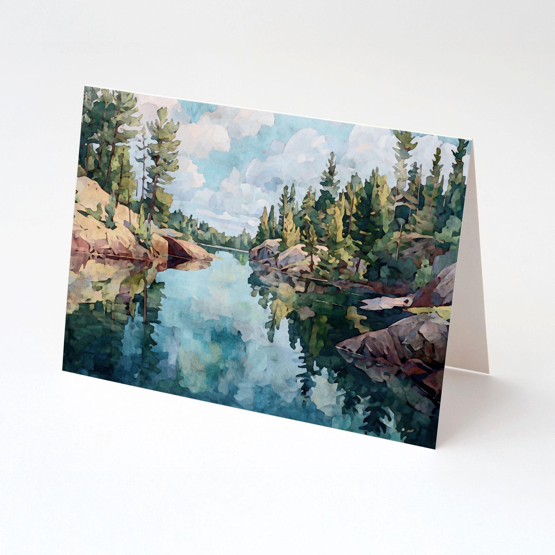 Folded greeting card featuring a watercolor-style painting of a peaceful northern lake, with rocky shores and tall evergreen trees reflected in the calm blue water under a partly cloudy sky. Displayed upright on a white background.