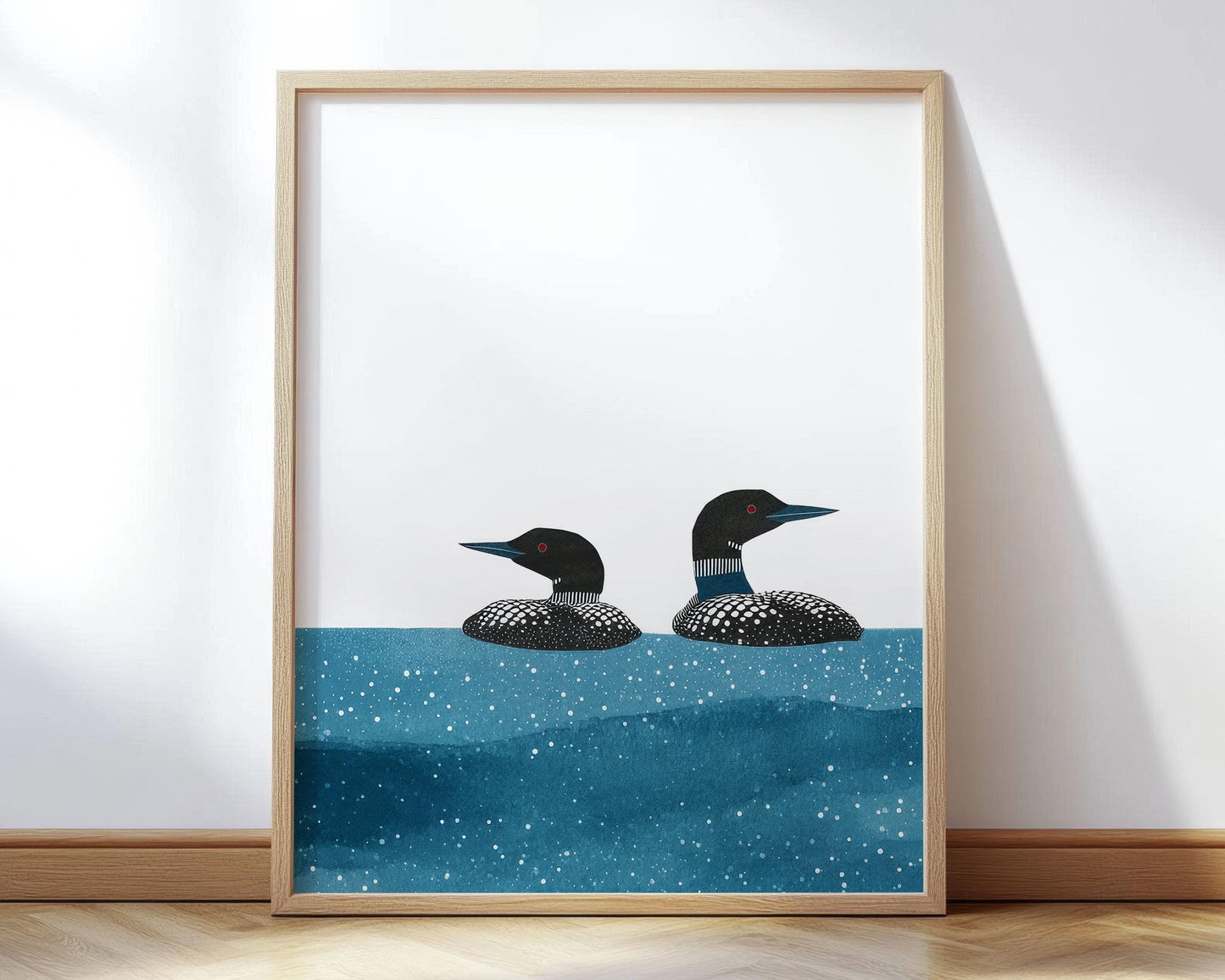 Framed vertical illustration of two loons with black-and-white patterned feathers and red eyes, floating in deep blue water speckled with white dots. The birds are shown against a clean white background, the artwork leaning against a wall on a light wood floor with natural sunlight casting soft shadows.