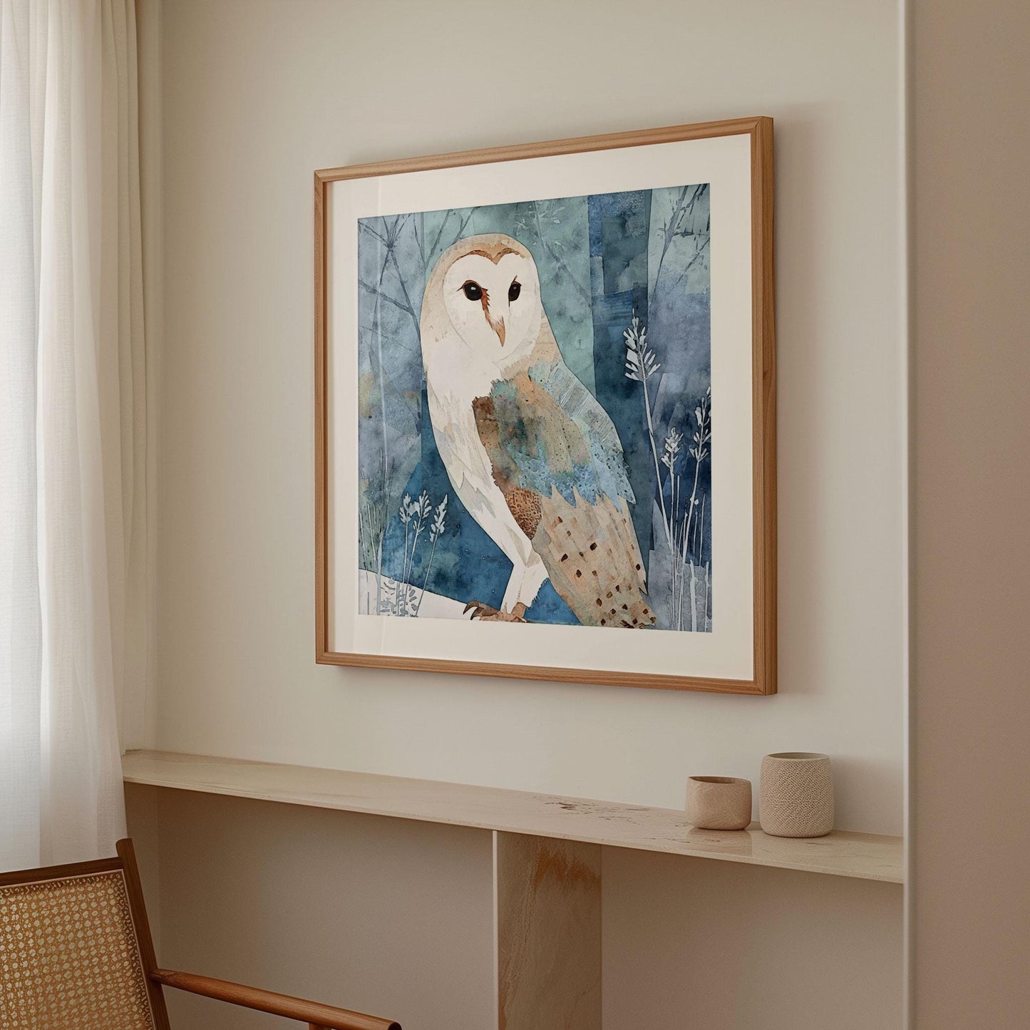 Framed watercolor-style barn owl print displayed on a light beige wall above a narrow wooden shelf. The artwork shows a soft, abstract mix of blue, teal, and tan tones. Natural light filters through white curtains beside a wooden chair, creating a warm, minimalist, and serene interior setting.