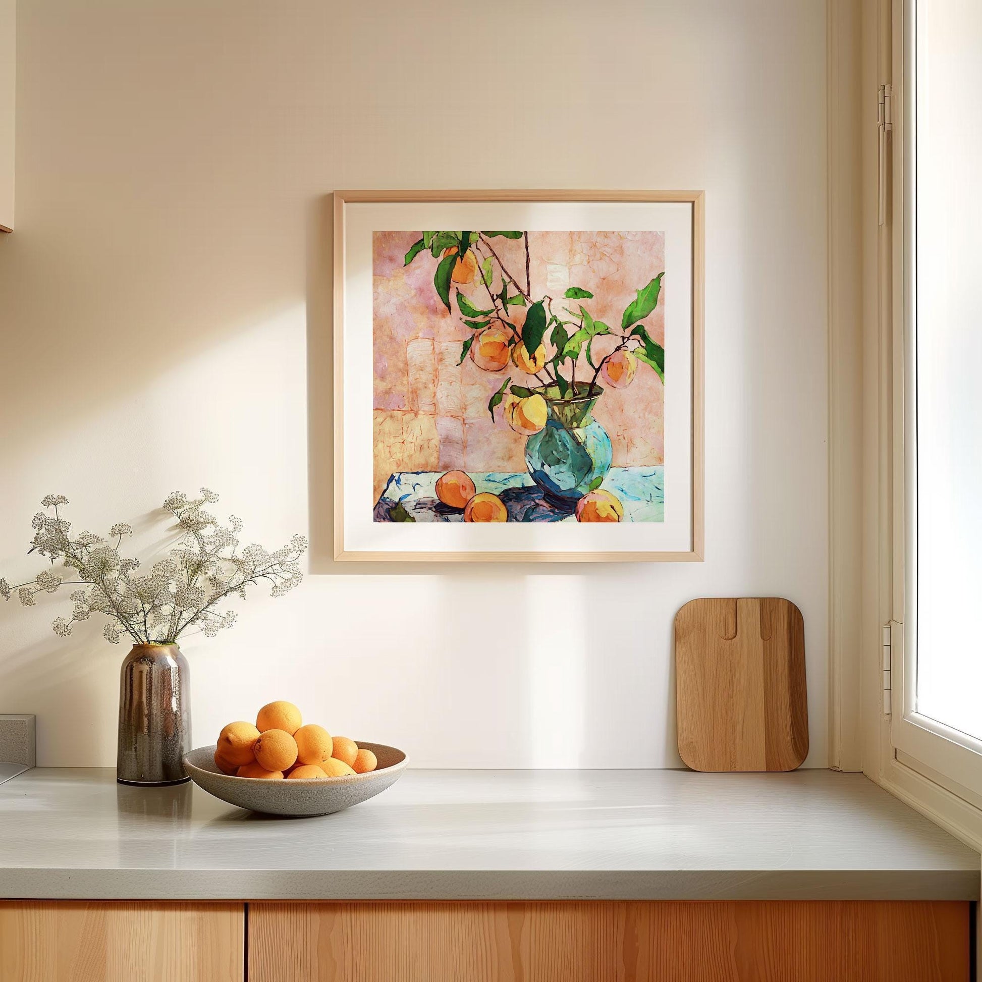 Square-framed still life painting of peaches in a blue glass vase, hung on a light kitchen wall near a window. The artwork shows leafy branches with ripe peaches against a textured pink background. Below, a countertop is styled with a ceramic vase of delicate white flowers, a wooden cutting board, and a bowl filled with fresh oranges, creating a bright and inviting scene.