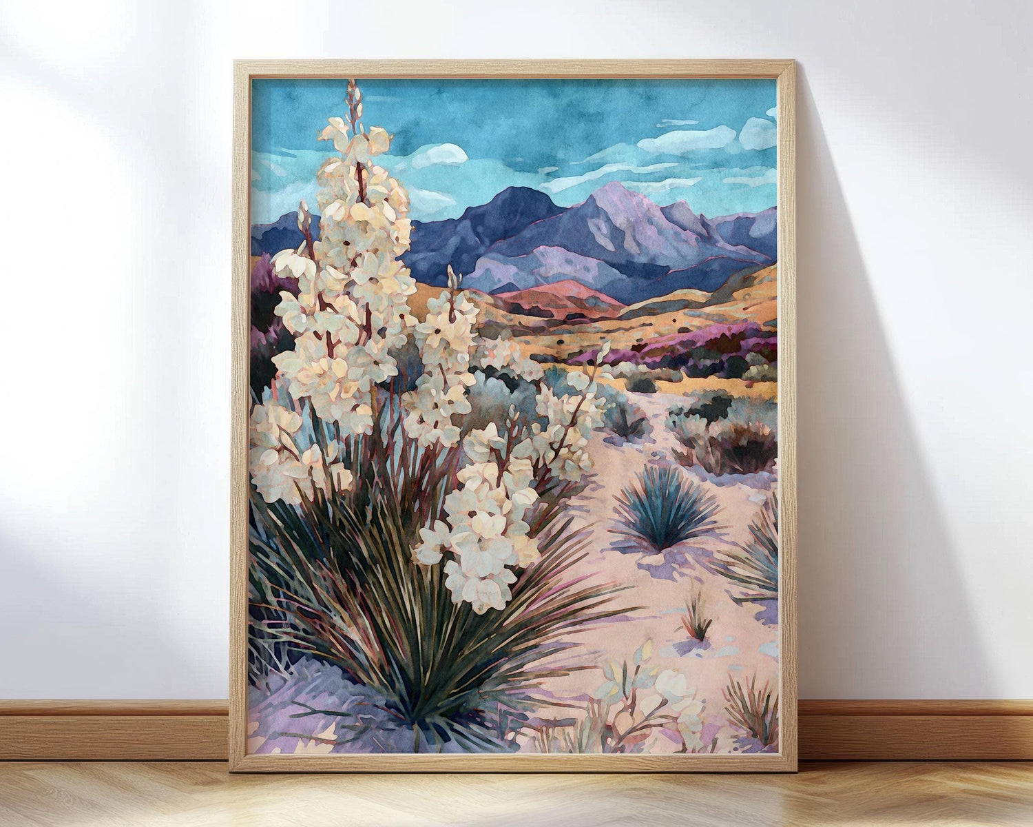 Framed watercolor-style desert painting featuring blooming yucca plants in the foreground, rolling sandy hills with shrubs, and purple-hued mountains beneath a vivid blue sky with soft clouds.